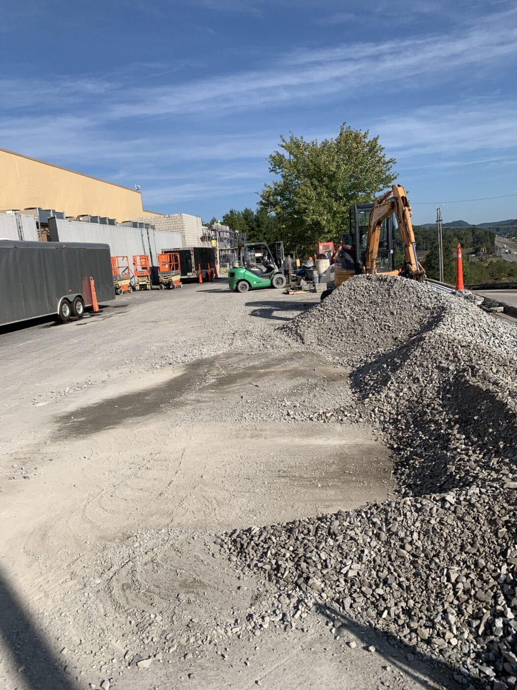 Construction site with a pile of gravel, an excavator, and various construction vehicles near a building. Safety cones line the area. Clear blue sky in the background.