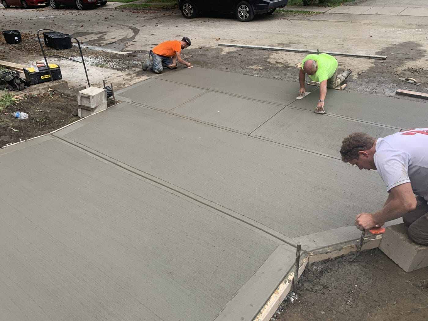 Three workers in safety vests smoothing wet concrete on a driveway using tools.