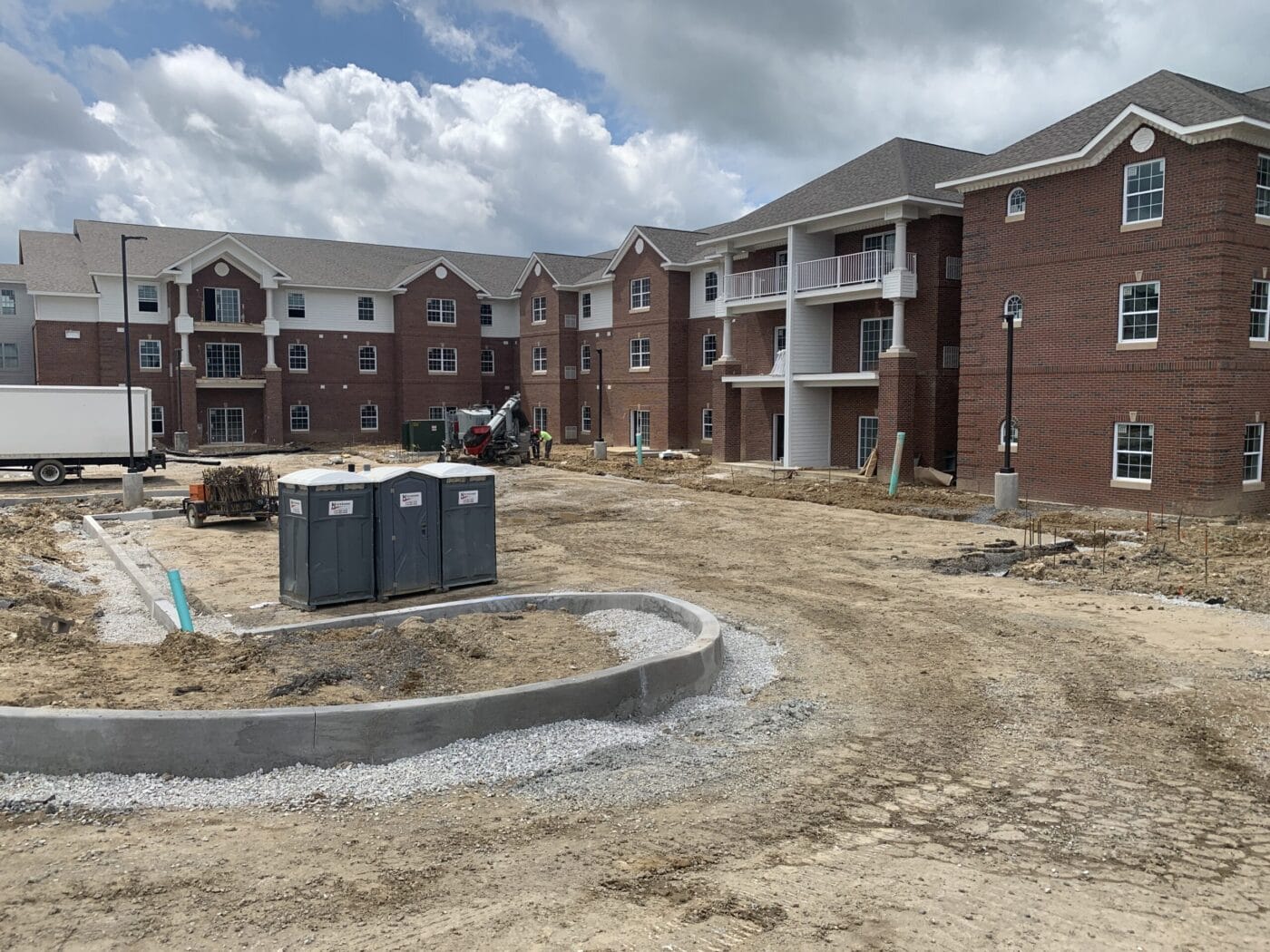 A partially completed three-story building with red brick exterior, surrounded by construction materials and portable toilets under a cloudy sky.