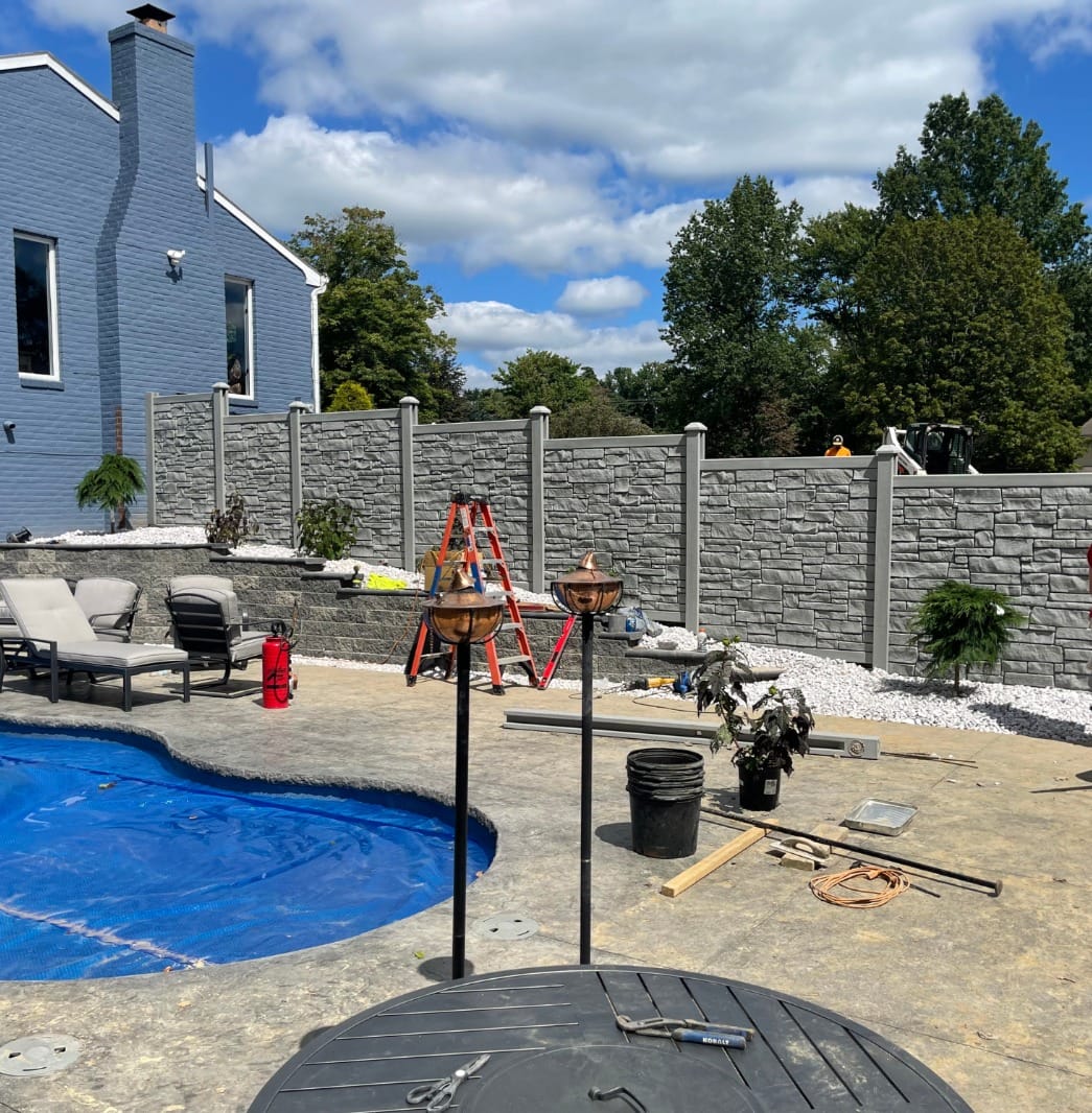 Backyard with a blue-covered pool, construction tools, ladders, and materials scattered. Grey stone fence in the background and a blue house to the left under a partly cloudy sky.