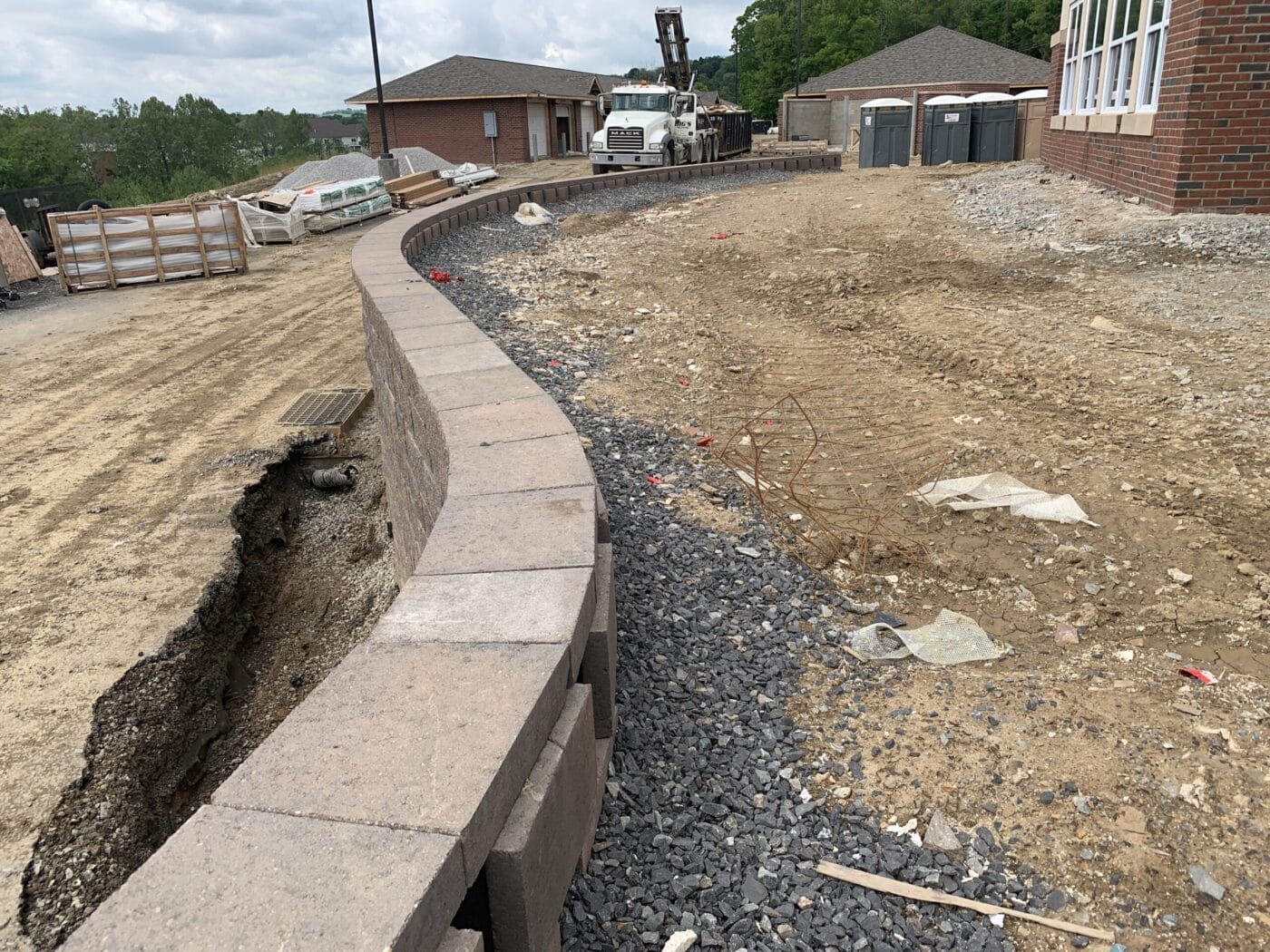 Construction site with a curved retaining wall, gravel, and dirt. A truck and portable toilets are in the background near brick buildings.