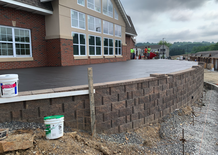 A newly built brick retaining wall supports a concrete patio in front of a large building. Construction materials are visible, and workers are gathered in the distance.