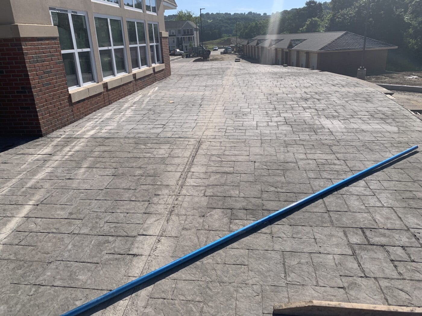 Concrete walkway with a textured surface alongside a brick building and several smaller structures in the distance under a clear sky. A blue pole lies across the foreground.