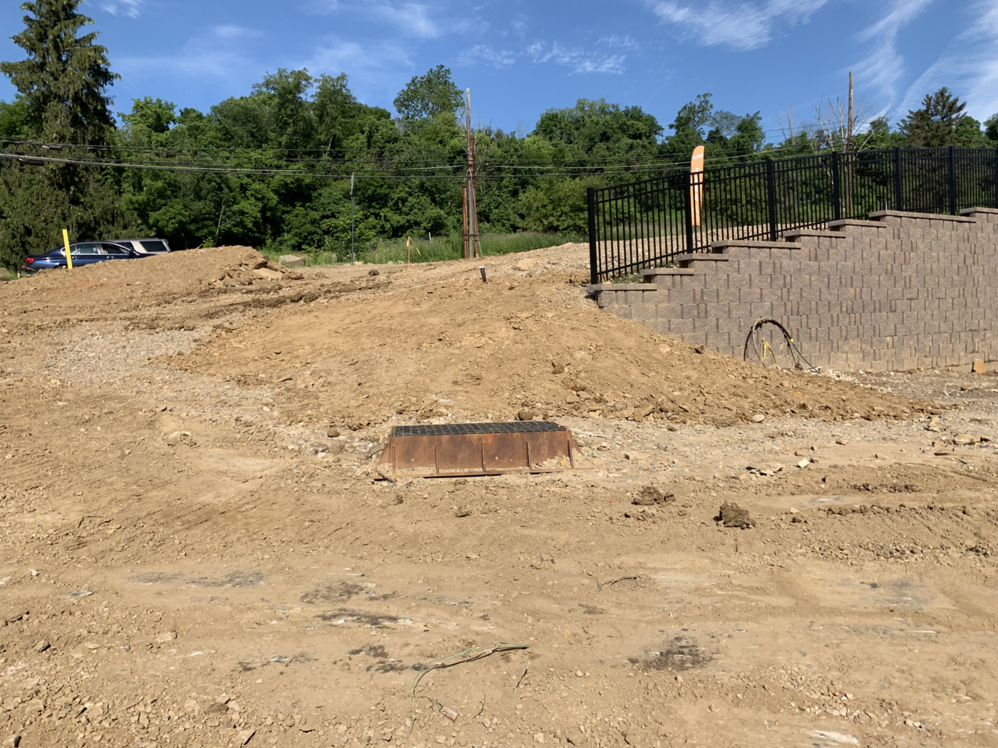 A dirt slope with a partially built retaining wall and a metal box in the foreground. Trees and parked cars are visible in the background under a blue sky.