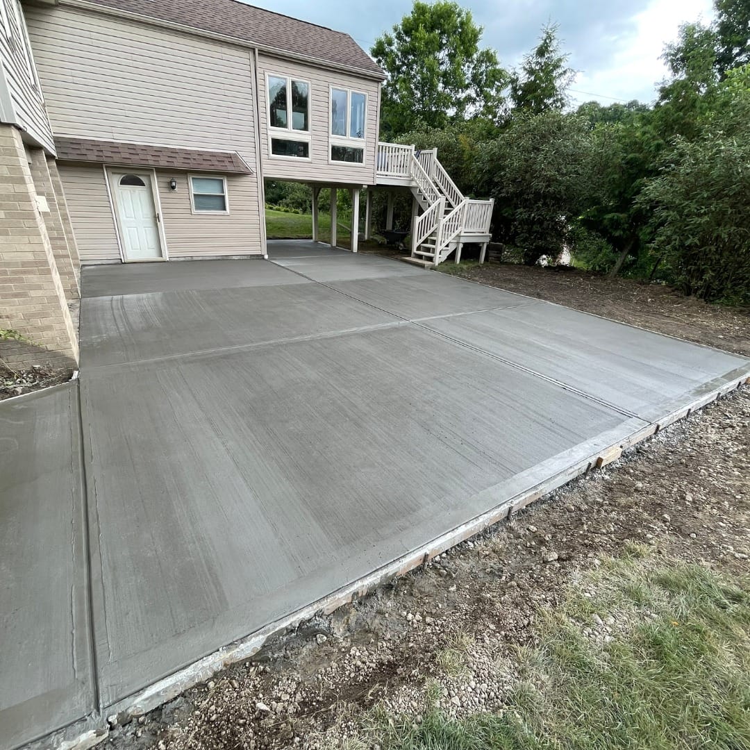 Freshly poured concrete patio adjacent to a two-story house with a balcony and staircase.