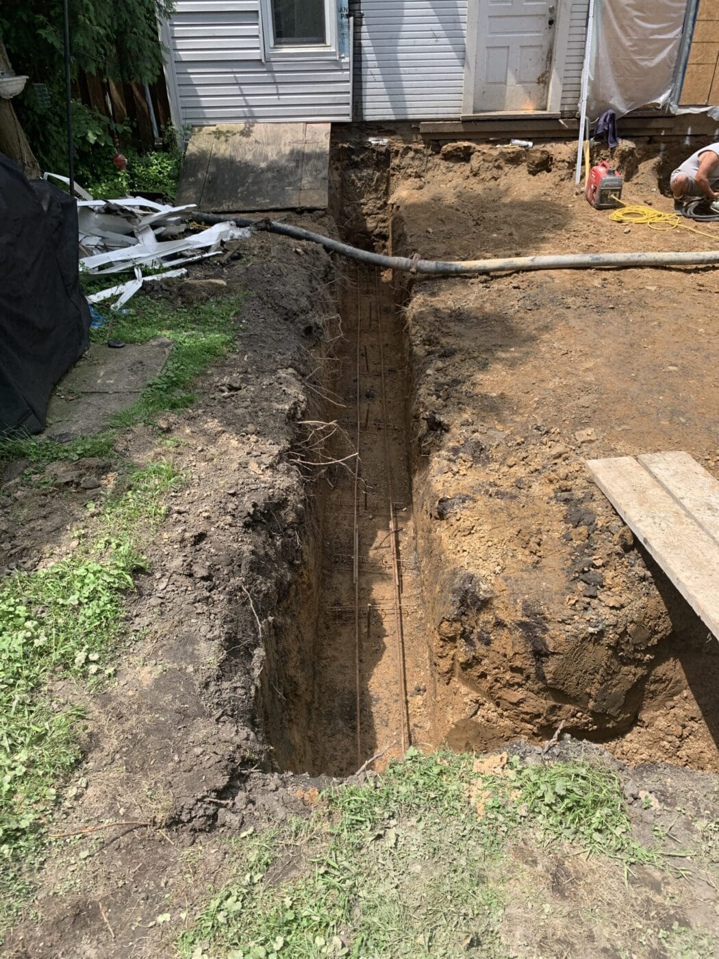 Deep trench dug in backyard, with exposed soil and visible reinforcing bars. A house is in the background. Construction materials and equipment are nearby.