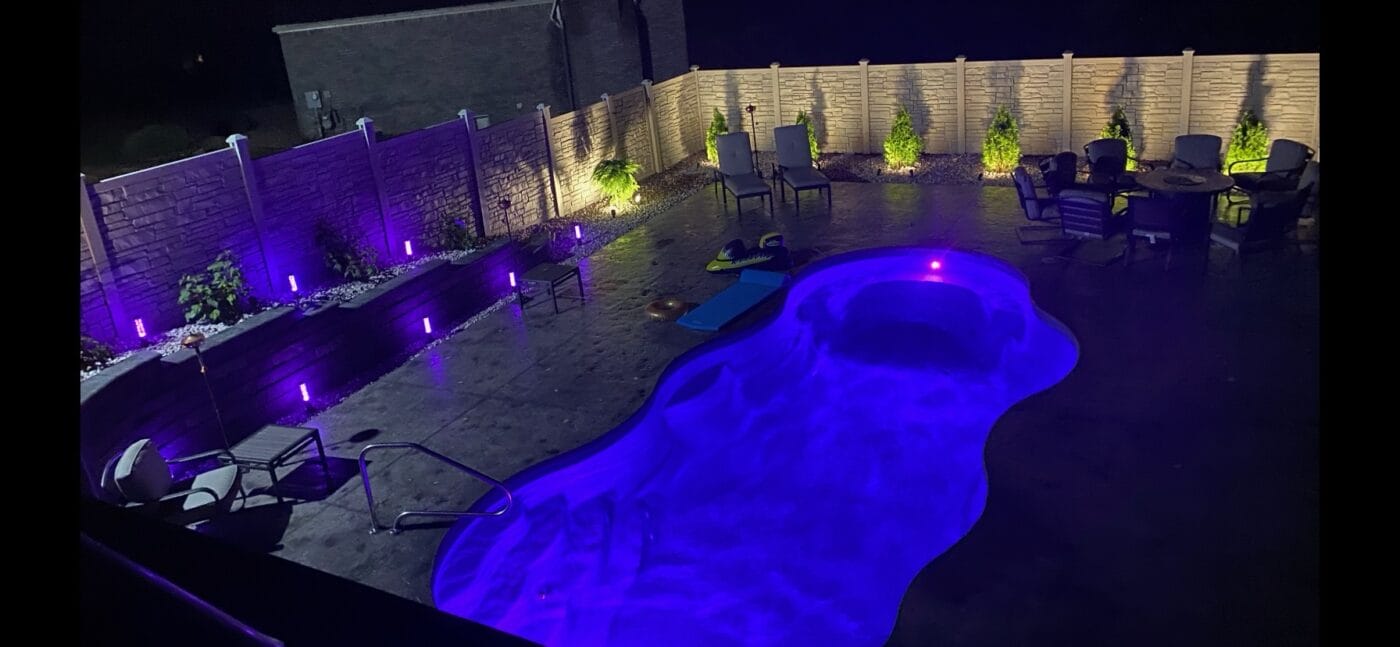 A backyard pool illuminated with purple lights at night, surrounded by chairs and a privacy fence, creating a serene atmosphere.